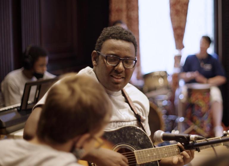 ABLE Day Session student strums a guitar and wears a gray Berklee t-shirt