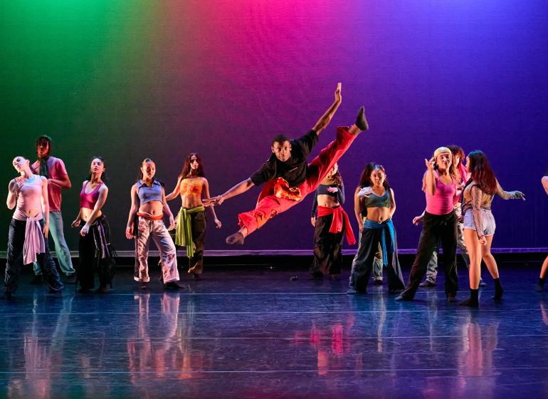 A group of dancers on stage performing a dynamic routine. One dancer in the center leaps high into the air with one leg extended and one arm reaching upward, wearing vibrant red pants and a black shirt. The other dancers are spread across the stage, each striking expressive poses and wearing colorful, casual dance attire. The background lighting is a gradient of vivid colors, including green, pink, and blue, creating an energetic and vibrant atmosphere.